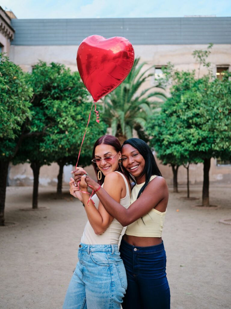 Loving lesbian couple posing together outdoors with a red heart-shaped balloon.